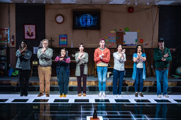 Eight people stand in a straight line on a reflective, grid-patterned floor, all with their arms crossed in front of their chests in synchronized gestures. They are dressed in casual clothing and appear focused, with varied facial expressions. Behind them is a church basement wall with a piano, clock, cross, bulletin boards, and children’s decorations. A metronome sits on the floor in front of them.