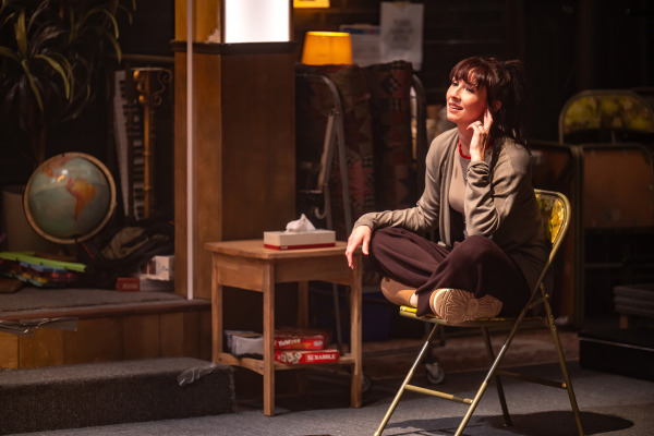 A woman sits cross-legged on a folding chair, smiling with one hand resting near her ear as if listening. She wears relaxed, casual clothing and is bathed in warm lighting. Beside her is a small wooden table with a tissue box on top and board games stored underneath, including Scrabble and Yahtzee. In the background of the church basement setting are stacked chairs, a globe, and various supplies.