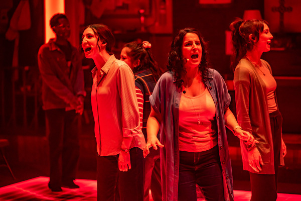 Four women stand in a row under intense red lighting, singing with expressive faces and emotional energy. They wear casual clothing and headset microphones. Two other figures are visible in the background, partially obscured and in softer focus. The setting is a church basement, with elements like a cross on the wall, a piano, and a bulletin board faintly visible in the dim red glow.