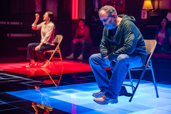 A man sits on a folding chair with his head lowered and hands clasped, appearing deep in thought, lit by blue light on a reflective, grid-patterned floor. Behind him, a woman sits on another chair under red lighting, holding one hand in the air as if making a point. Other people are visible in the dimly lit background of the church basement, including a person seated near a lit lamp and a piano.