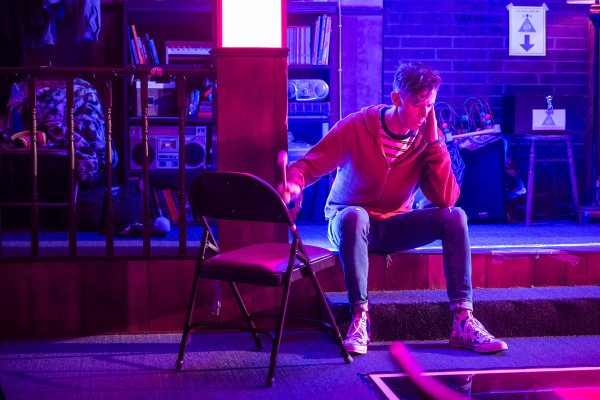 A person sits on the edge of a raised platform in a church basement, resting their head on one hand and holding a mallet in the other. They wear a red hoodie, striped shirt, rolled-up jeans, and patterned high-top sneakers. The space is lit in vibrant pink and purple tones. Behind them are children’s toys, books, a boombox, and a sign with a large arrow pointing down. A folded blanket and other personal items are visible on a nearby chair.