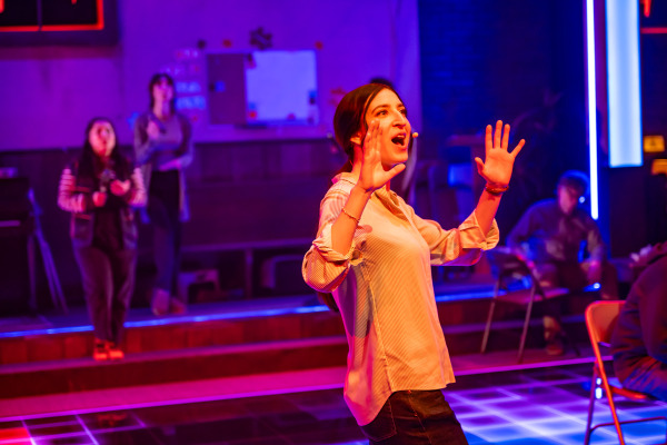 A woman in the foreground sings with her hands raised and expressive face, illuminated by vibrant purple and red stage lighting. Two other people stand in the background near the steps of a stage area, slightly out of focus. The setting is a church basement, with folding chairs, a bulletin board, and wall decorations partially visible in the dim light.