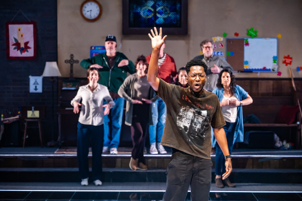 A man stands at the front singing with one arm raised and an expressive face. Behind him, seven other people stand in a staggered formation with their hands positioned in front of their chests, facing forward. The setting is a church basement with a piano, bulletin board, clock, and colourful decorations visible on the back wall.