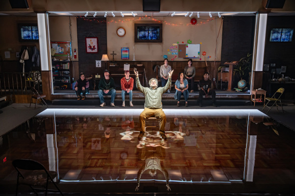 A man sits alone at the centre of a highly reflective floor with his arms raised and face lifted, framed by a glowing square of light. Behind him, eight people sit in a row on risers against the wall of a church basement. The background includes a piano, bulletin boards, religious decorations, a globe, and various signs and crafts, creating a warm, lived-in environment. The man’s reflection is clearly visible on the glossy floor beneath him.