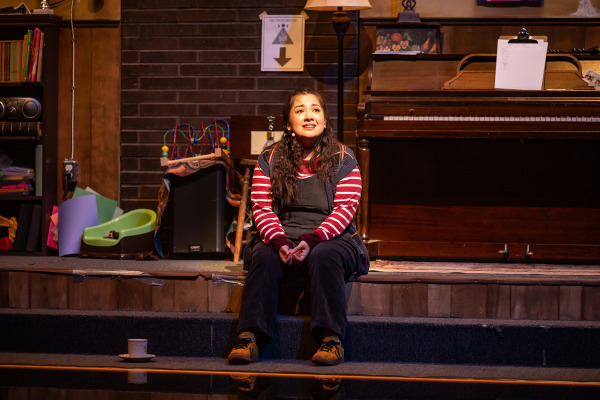 A woman sits on a step in front of a piano, looking upward with a thoughtful expression. She wears a striped long-sleeve shirt under dark overalls and yellow shoes. The setting is a church basement, with items such as a children's toy, bookshelf, lamp, and a cup and saucer on the floor. A clipboard and papers rest on the piano, and various decorations and signs are visible in the background.