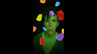 A man illuminated by green lighting stands against a dark background, looking directly at the camera with a neutral expression. Colourful gummy bears in red, yellow, orange, blue, and purple appear digitally overlaid and float around his face. The image has a vertical format with black bars on either side.