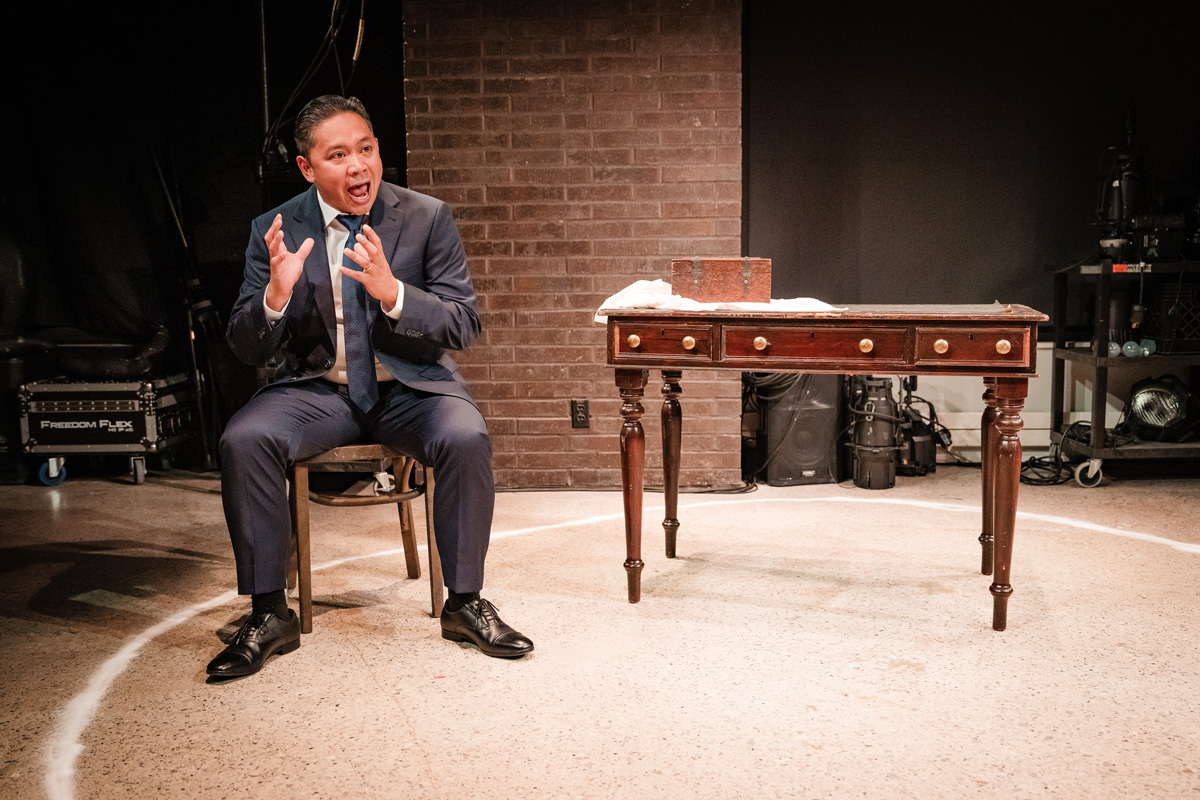 A man stands inside a chalk circle on the stage, leaning forward slightly with hands together in a pleading gesture. His jacket is draped over the back of a nearby chair. The wooden desk with a small box is to his side.