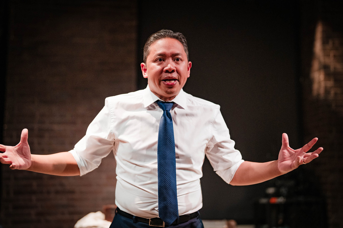 A man in a blue suit sits on a chair within a chalk circle on a stage, gesturing expressively with both hands and mouth open as if speaking. A wooden desk with drawers, a white cloth, and a small box is beside him. The background includes a brick wall and stage equipment.
