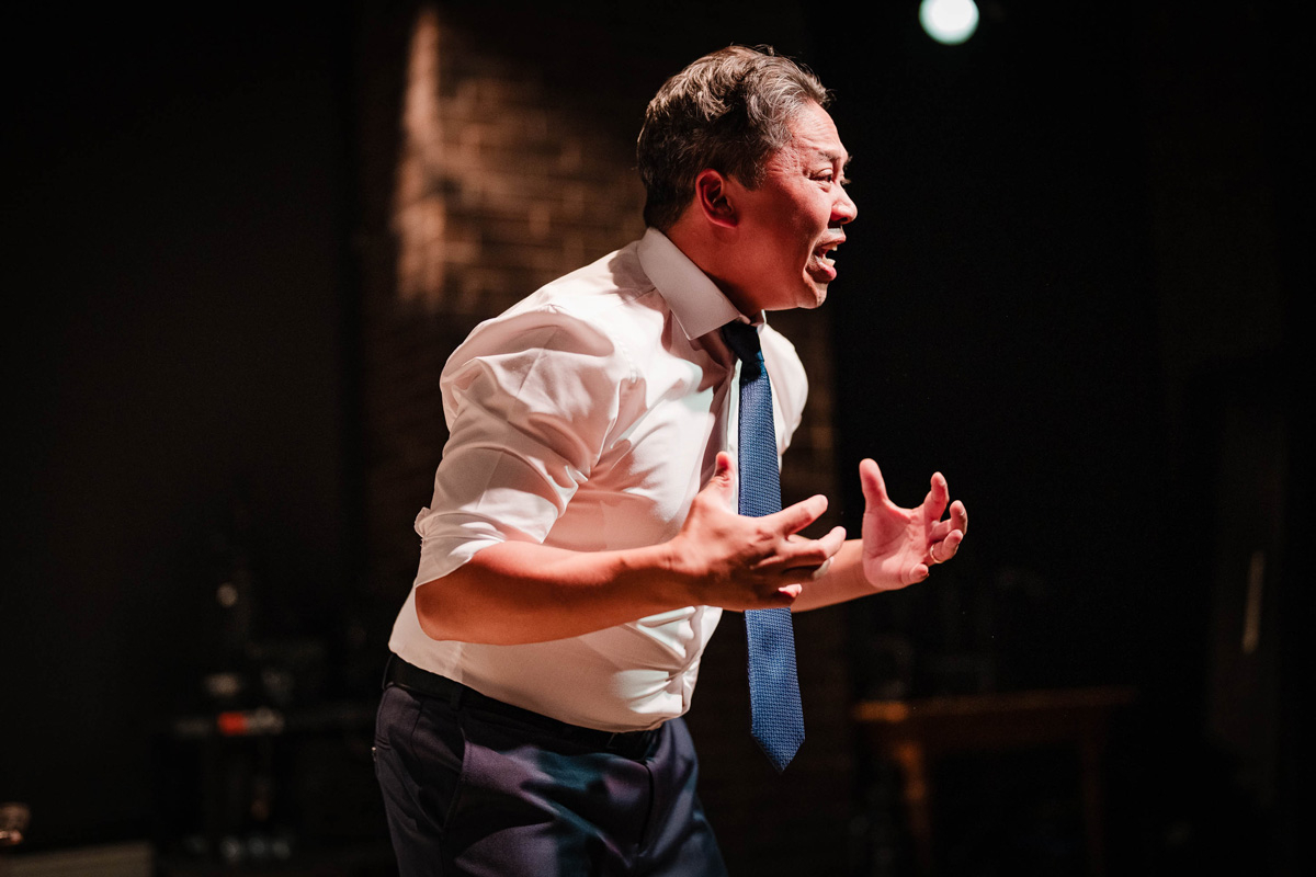 A man with rolled-up sleeves and a blue tie leans forward mid-speech with an intense expression, hands held out in front of him with fingers spread in a gripping gesture. He is lit dramatically against a dark background with a brick wall and some indistinct stage equipment.