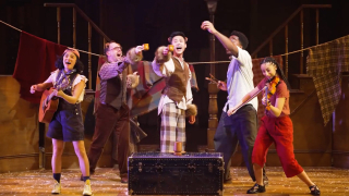 Five performers on a wooden stage are mid-performance, with joyful expressions and lively body language. From left to right: a woman in overalls and a pink cap plays an acoustic guitar and sings; a man in a vest and glasses raises a small cup, wearing furry cuffs and ears; a man in a brown plaid vest and pants, also with furry cuffs and ears, stands on a black trunk and holds a cup while singing; another man in a short-sleeved shirt and dark trousers raises his arm; and a woman in red pants plays a violin energetically. Behind them are wooden stairs, hanging blankets, and scattered white confetti on the floor.