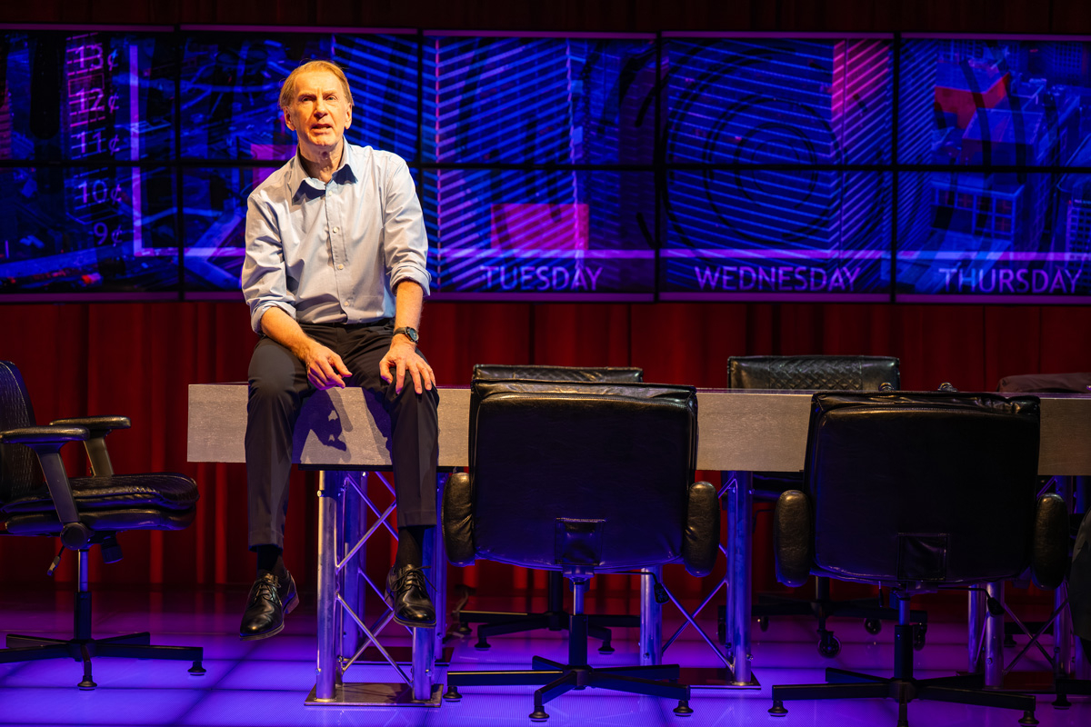 A man sits on a modern table at the centre of a stage, surrounded by black office chairs. Behind him is a wall of digital screens with vivid blue and purple graphics and the words 