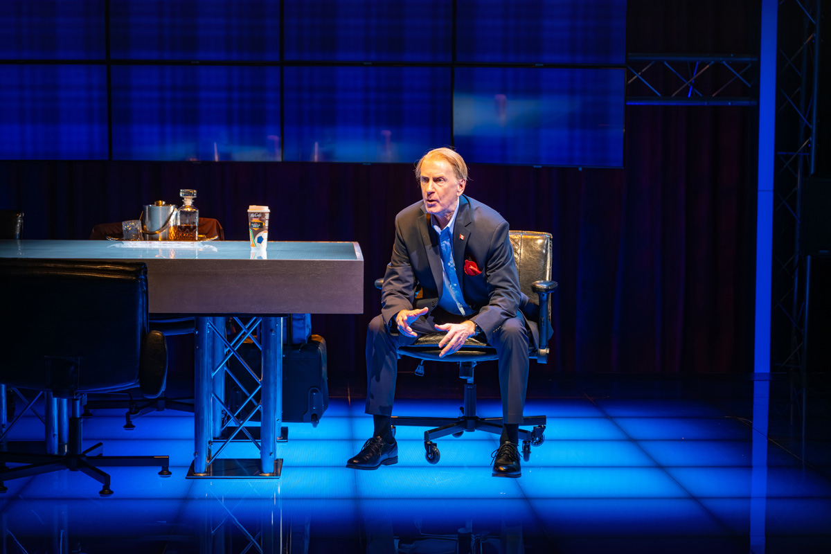 A man in a navy suit with a red pocket square sits on a black office chair at the end of a table, speaking expressively. The table holds a takeout coffee cup, a decanter of amber liquid, and glassware. The background is lit in deep blue tones, with a wall of digital screens.