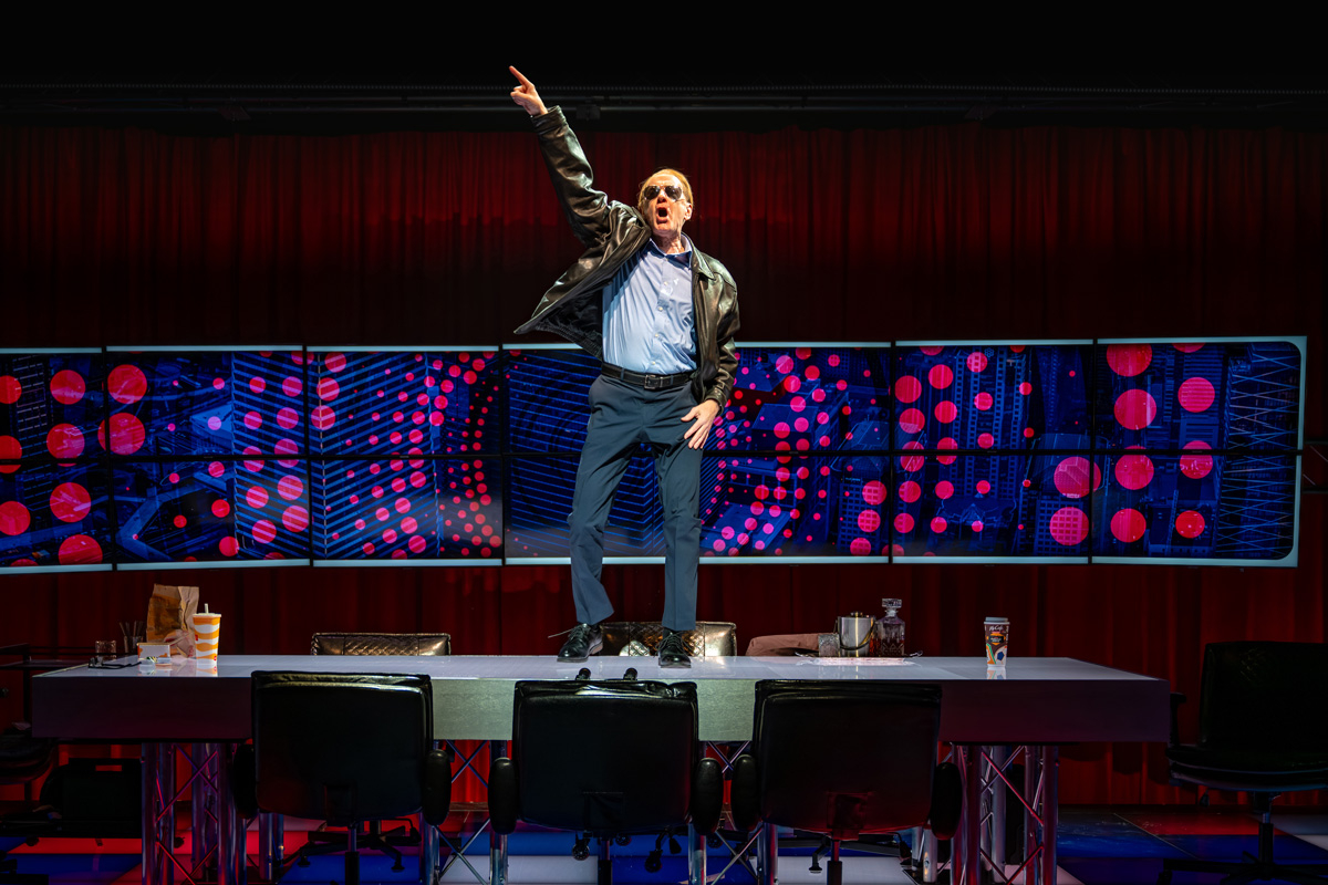 A man in a leather jacket, dress shirt, and slacks stands on top of a conference table, striking a dynamic pose with one arm pointing upward. The stage is lit with dramatic red and blue lighting, and the digital screen behind him shows a pattern of red circles on a blue background. Office chairs surround the table, and drinks are scattered across it.