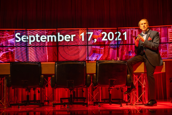 A man in a navy suit and red pocket square stands beside a long table with black office chairs, one leg resting on the table. Behind him, a wall of screens displays the date “September 17, 2021” in large white text over red-toned cityscape imagery.