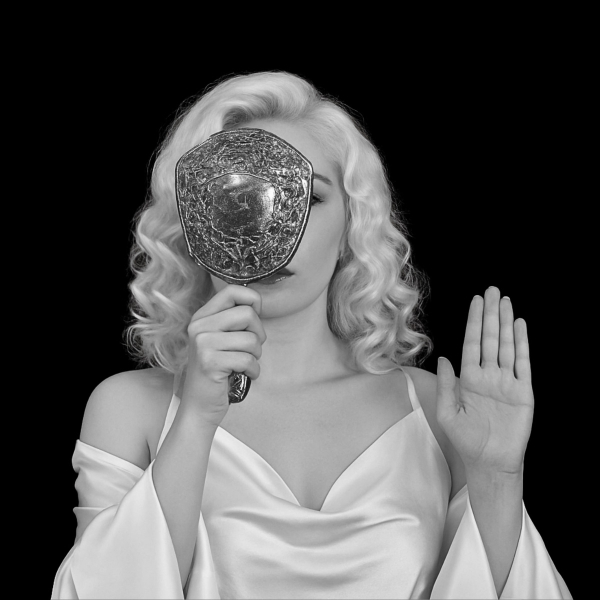 A woman with light, wavy, shoulder-length blonde hair is dressed in a satin, off-the-shoulder white gown. She holds an ornate, embossed hand mirror that covers one eye and part of her face. Her other hand is raised with the palm facing outward. The background is solid black, creating a high contrast with the subject and enhancing the monochrome aesthetic.