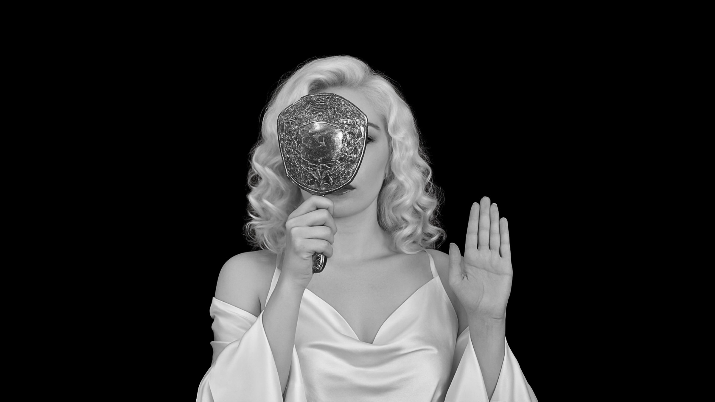 A woman with light, wavy, shoulder-length blonde hair is dressed in a satin, off-the-shoulder white gown. She holds an ornate, embossed hand mirror that covers one eye and part of her face. Her other hand is raised with the palm facing outward. The background is solid black, creating a high contrast with the subject and enhancing the monochrome aesthetic.