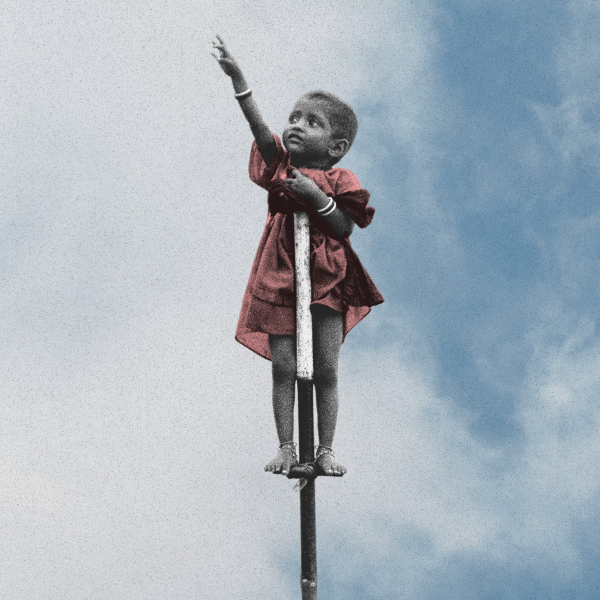 A small child in a red dress stands balanced on top of a narrow pole against a bright blue sky with large white clouds. The child is shown in black and white except for the dress, with one arm stretched upward and the other hand resting on their chest. Bracelets are visible on both wrists, and the child is barefoot, standing on a small support near the top of the pole.