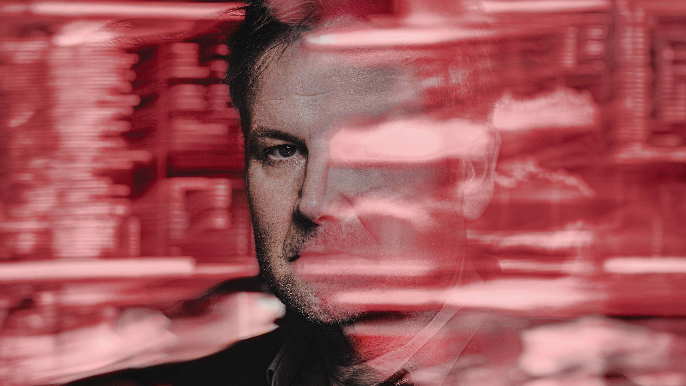 Close-up portrait of a man’s face partially obscured by horizontal red motion blur streaks, with one eye sharply visible and the background filled with blurred shelves and objects in red tones. He has short dark hair, light stubble, and a serious expression, and is wearing a dark jacket over a collared shirt.
