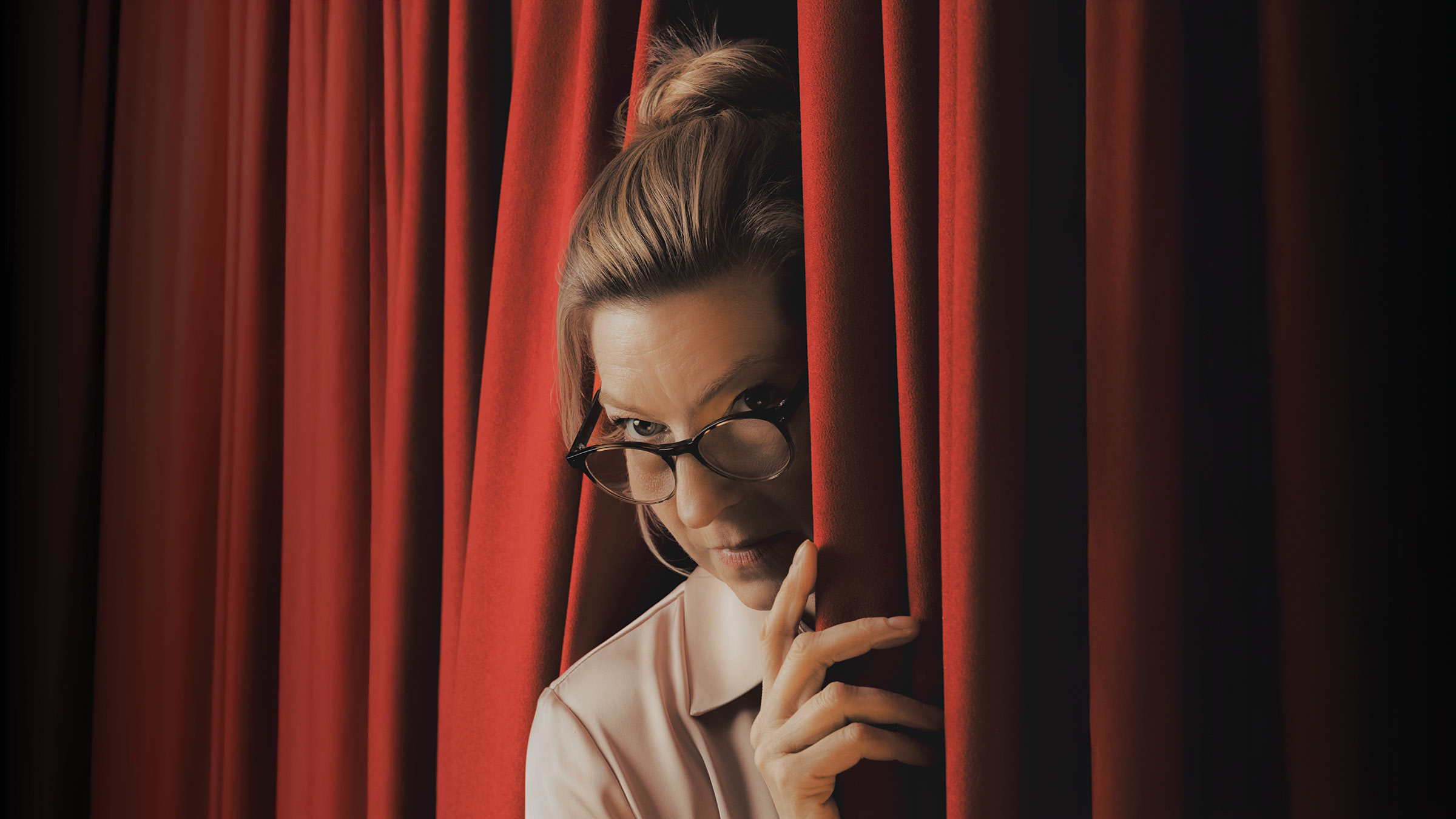 A woman with her hair in a loose bun peers through a narrow gap in rich red stage curtains. She wears dark-framed glasses low on her nose and a pale collared blouse, with one hand holding the curtain aside. Warm lighting highlights her face and hand against the dark folds of the curtain.
