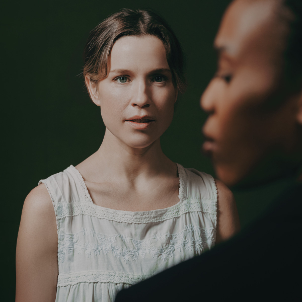 A woman in a sleeveless white dress looks forward with a calm, intent expression, while the blurred side profile of another person fills the right foreground. The woman’s hair is pulled back with loose strands framing her face, and the dark background is lit with soft green tones.