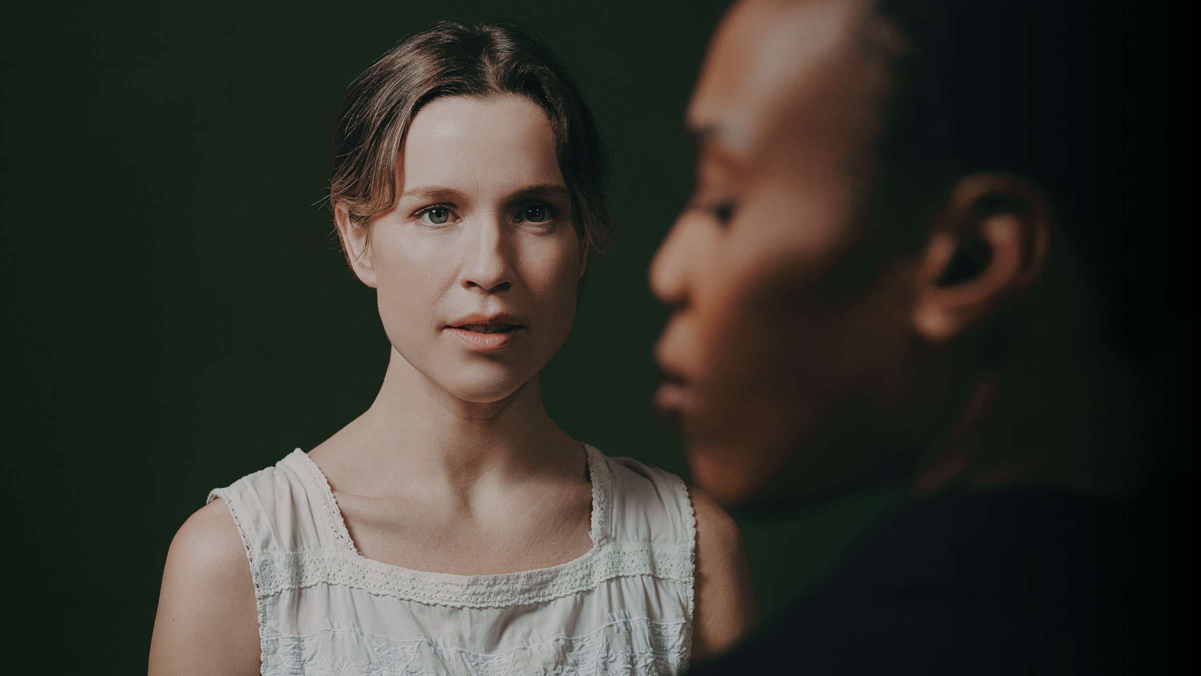 A woman in a sleeveless white dress looks forward with a calm, intent expression, while the blurred side profile of another person fills the right foreground. The woman’s hair is pulled back with loose strands framing her face, and the dark background is lit with soft green tones.