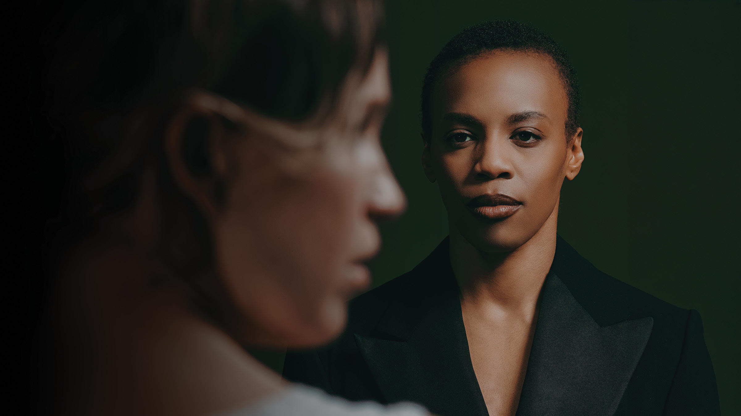 A sharply focused person with very short hair and a black jacket looks ahead with a calm, serious expression, while the blurred profile of another person in white fills the left foreground. The background is dark with soft green lighting.