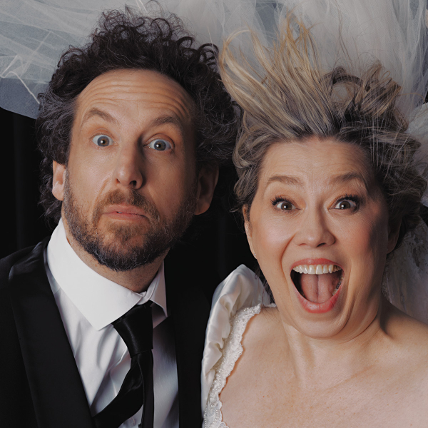 A man in a black suit and tie and a woman in a white wedding dress pose closely together against a dark background, both staring wide-eyed at the camera with surprised expressions. The woman’s mouth is open in a shout or laugh, and her blonde hair is blown upward beneath a sheer white veil that spreads across the top of the image. The man has curly dark hair and a beard, and the woman’s dress has an off-the-shoulder neckline with lace trim.
