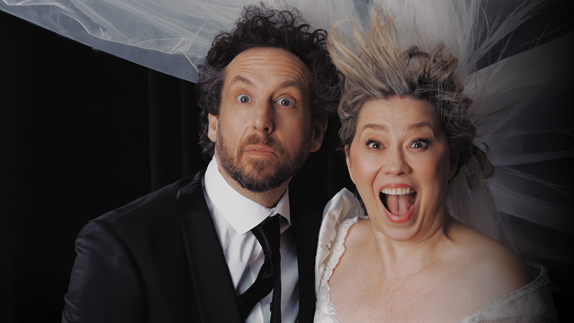 A man in a black suit and tie and a woman in a white wedding dress pose closely together against a dark background, both staring wide-eyed at the camera with surprised expressions. The woman’s mouth is open in a shout or laugh, and her blonde hair is blown upward beneath a sheer white veil that spreads across the top of the image. The man has curly dark hair and a beard, and the woman’s dress has an off-the-shoulder neckline with lace trim.