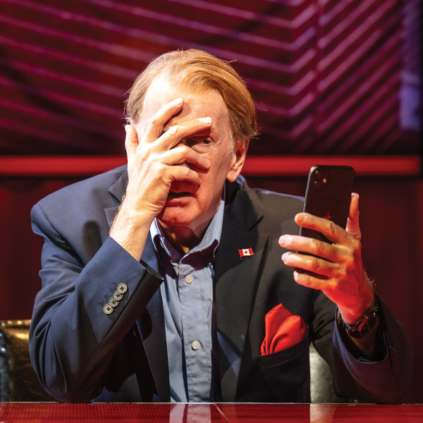 An older man in a navy blazer and light blue shirt sits at a glossy desk under red stage lighting, holding a smartphone in one hand while covering much of his face with the other. A small Canadian flag pin is attached to his lapel, and a red pocket square is tucked into his jacket. Large screens with red and purple geometric patterns fill the background.