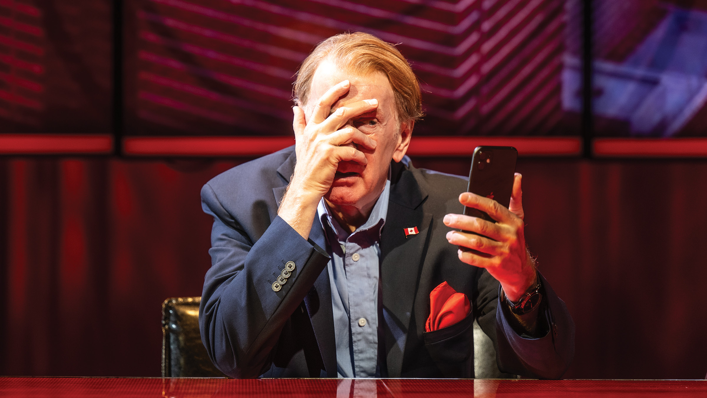 An older man in a navy blazer and light blue shirt sits at a glossy desk under red stage lighting, holding a smartphone in one hand while covering much of his face with the other. A small Canadian flag pin is attached to his lapel, and a red pocket square is tucked into his jacket. Large screens with red and purple geometric patterns fill the background.