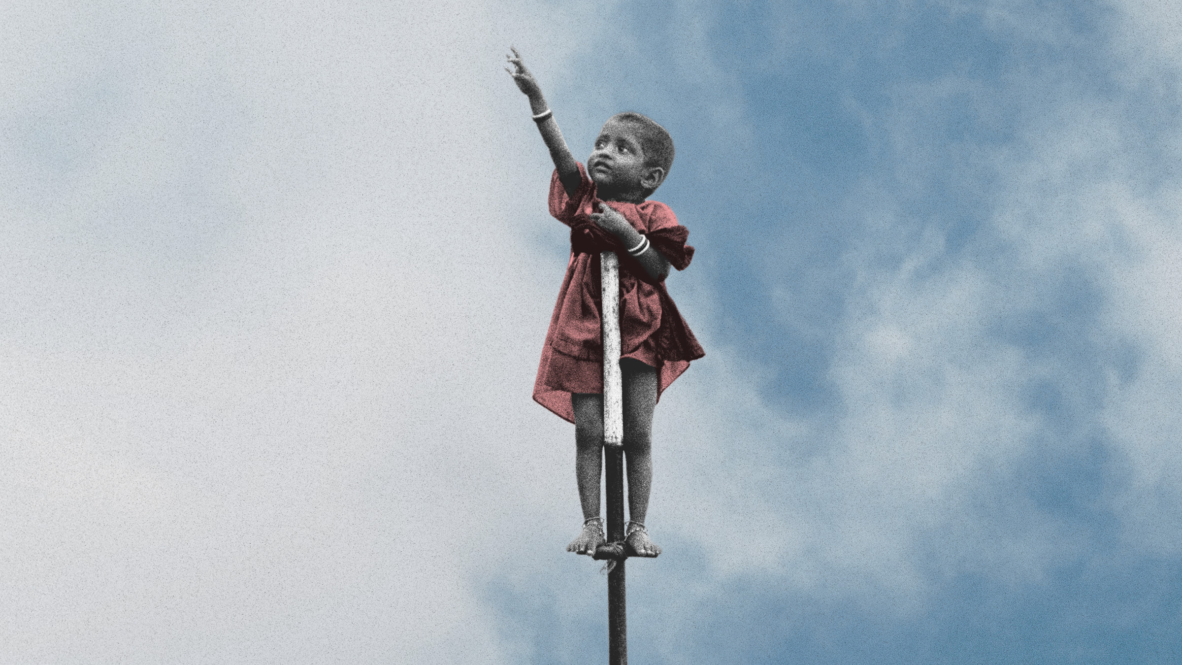 A small child in a red dress stands balanced on top of a narrow pole against a bright blue sky with large white clouds. The child is shown in black and white except for the dress, with one arm stretched upward and the other hand resting on their chest. Bracelets are visible on both wrists, and the child is barefoot, standing on a small support near the top of the pole.